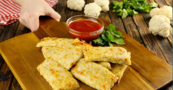 Einige Blumenkohl-Käse-Snacks auf einem Holzbrett. Daneben eine Schale mit Dip und Blumnkohlröschen im Hintergrund.