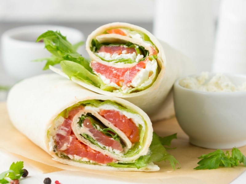 2 Wraps mit Lachs auf einem Brett, daneben frische Petersilie, Pfefferkörner und eine kleine Schüssel mit Frischkäse.