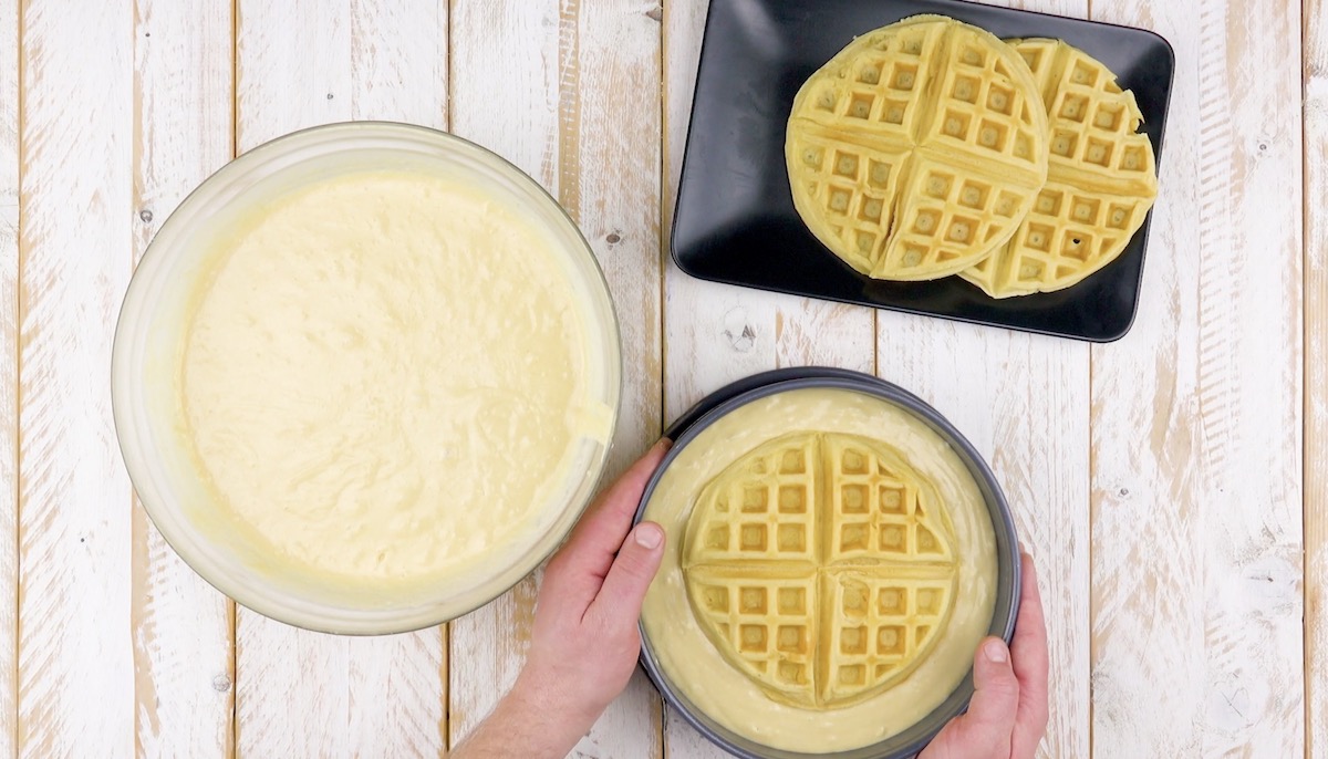 Eine Waffel wird auf flÃ¼ssigen Teig in Kuchenform gelegt, im Hintergrund zwei fertige Waffeln auf Teller und eine SchÃ¼ssel mit Teig daneben