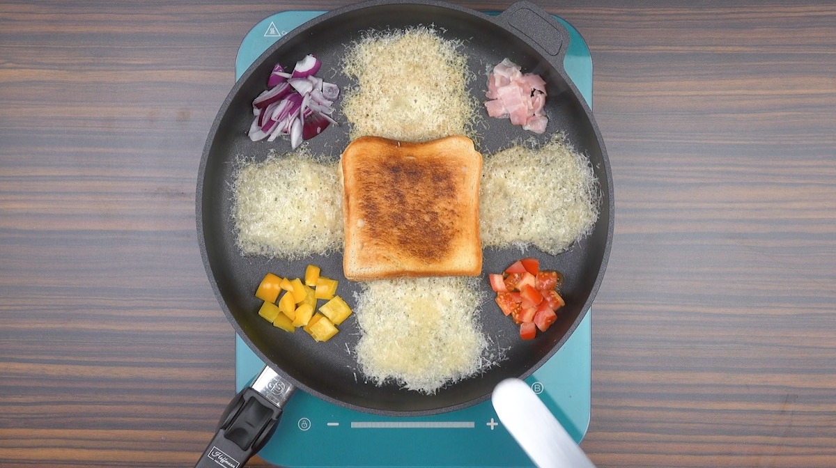 Eine Pfanne von oben, mit einem Kreuz aus Parmesan, Toastbrot in der Mitte und Paprika, Zwiebeln und Schinken in den freien Ecken