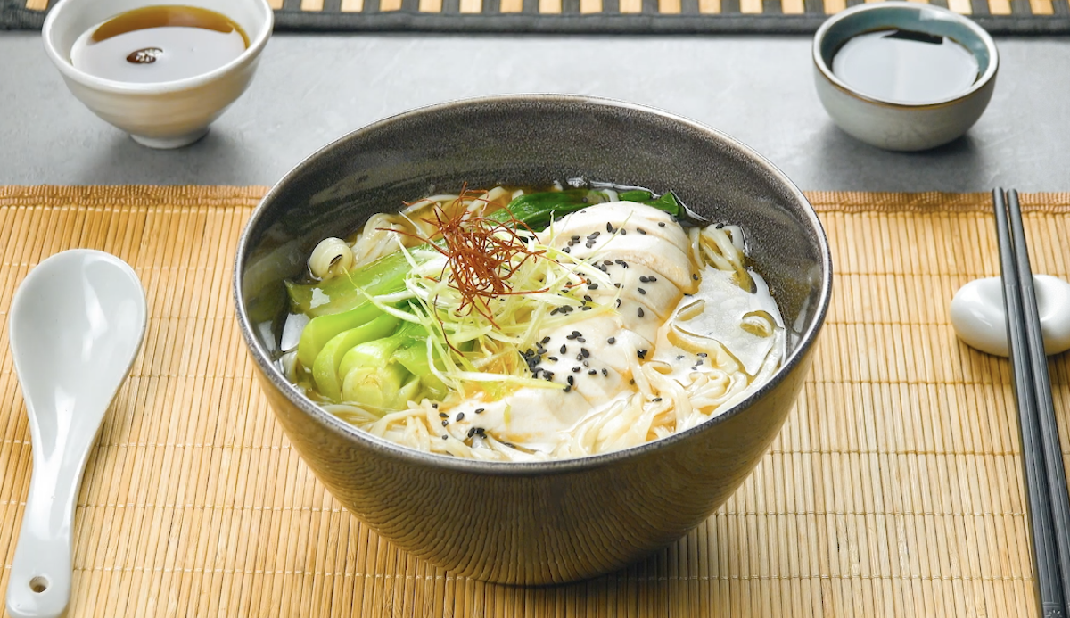 Ramen in einer Schüssel. Daneben stehen Schalen mit Soße und Besteck.