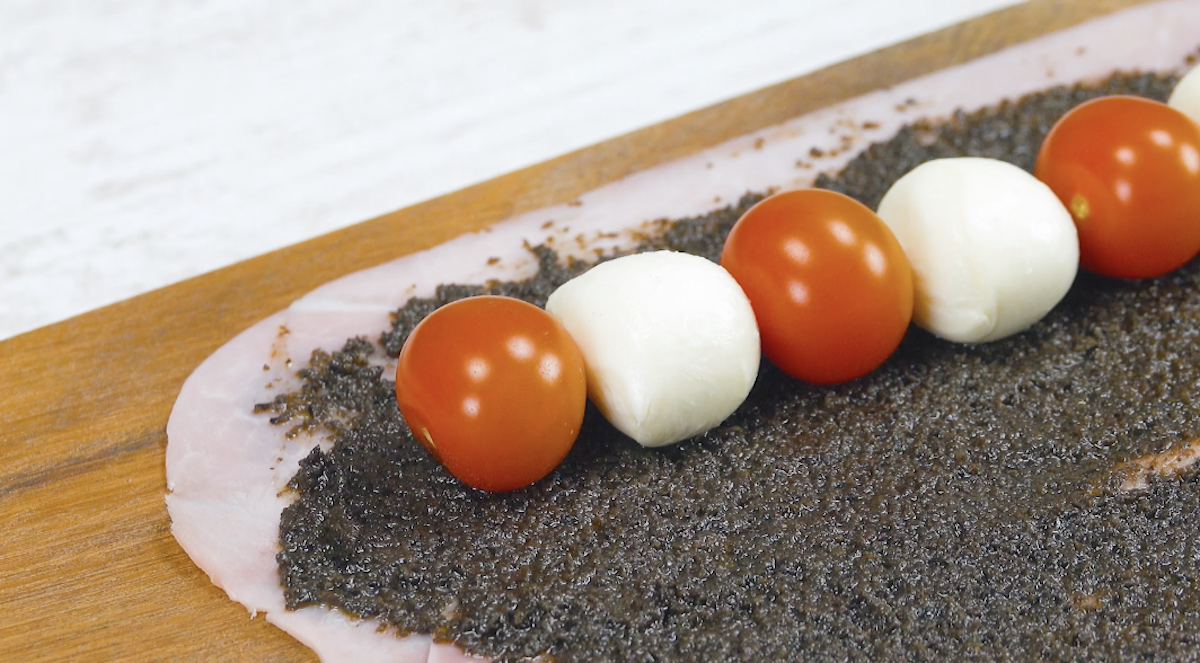 Kirschtomaten und Mini-Mozzarellas werden abwechselnd in Reihe auf Kochschinken mit Olivencreme gelegt