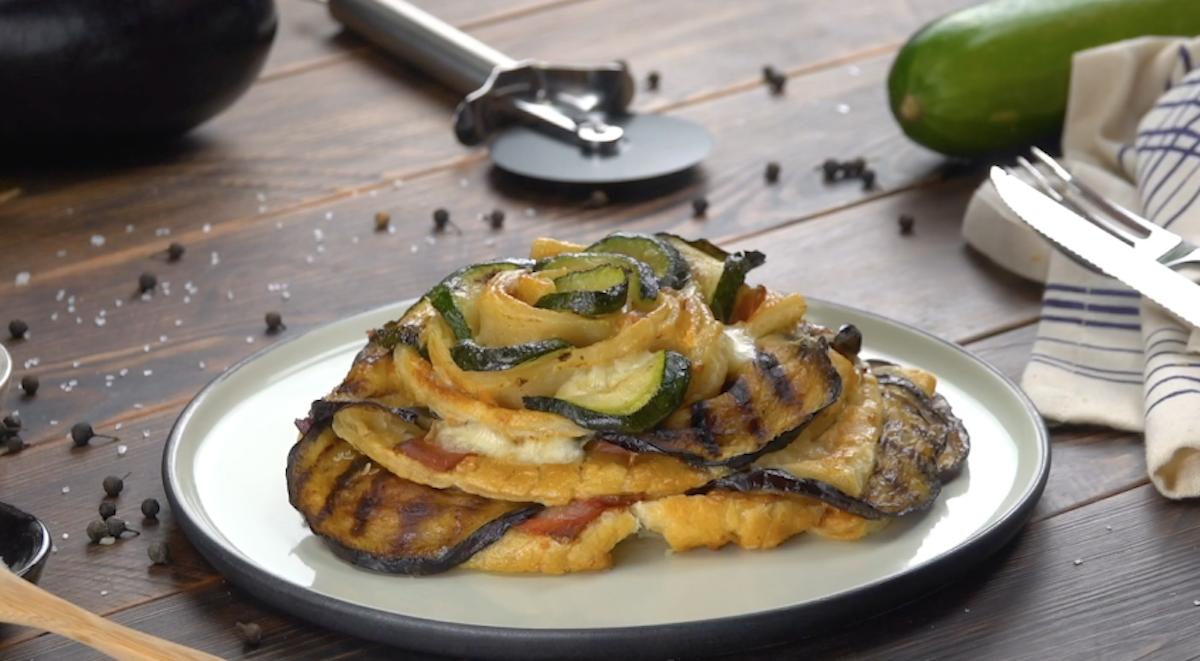 Eine Blätterteigrose mit Aubergine auf einem Teller. Im Hintergrund Besteck, eine Zucchini und eine Aubergine.