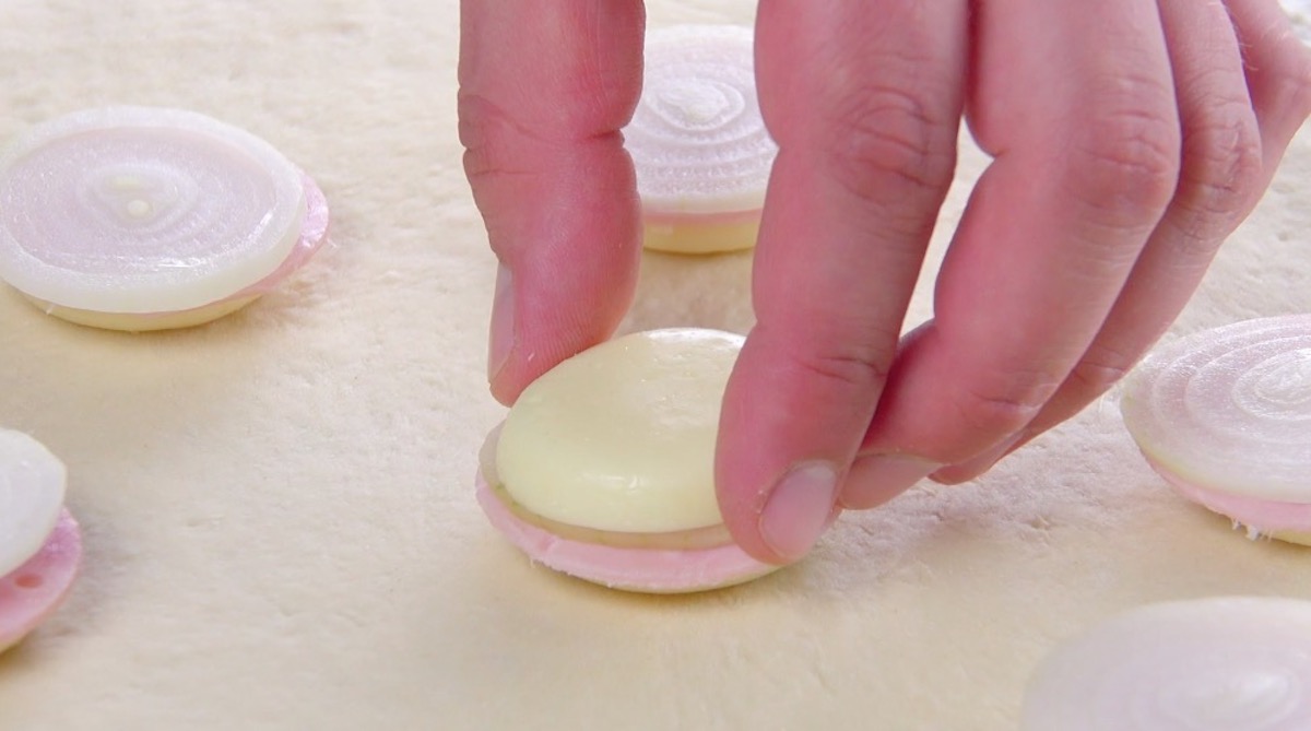 Halbierter Babybel-KÃ¤se wird auf Zwiebelring und Kochschinkenscheibe auf Pizzateig gelegt