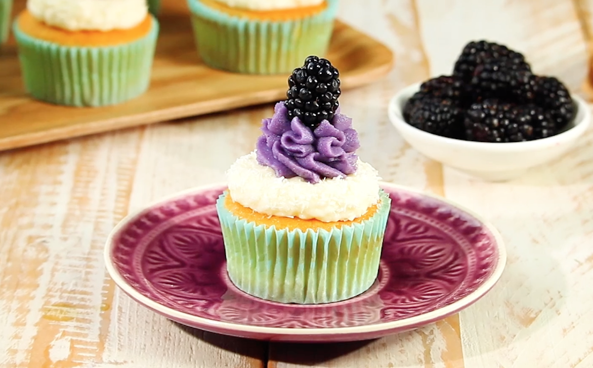 Ein Brombeer-Kokos-Muffin auf einem Teller. Daneben steht eine Schale mit frischen Brombeeren und weitere Muffins im Hintergrund.