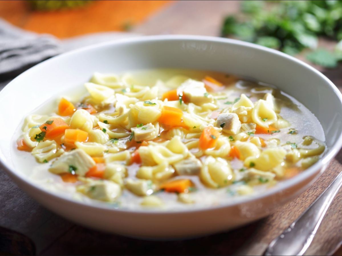 Ein Teller Hühnersuppe mit Nudeln. Daneben ein Löffel und ein Geschirrtuch. Im Hintergrund frische Petersilie.