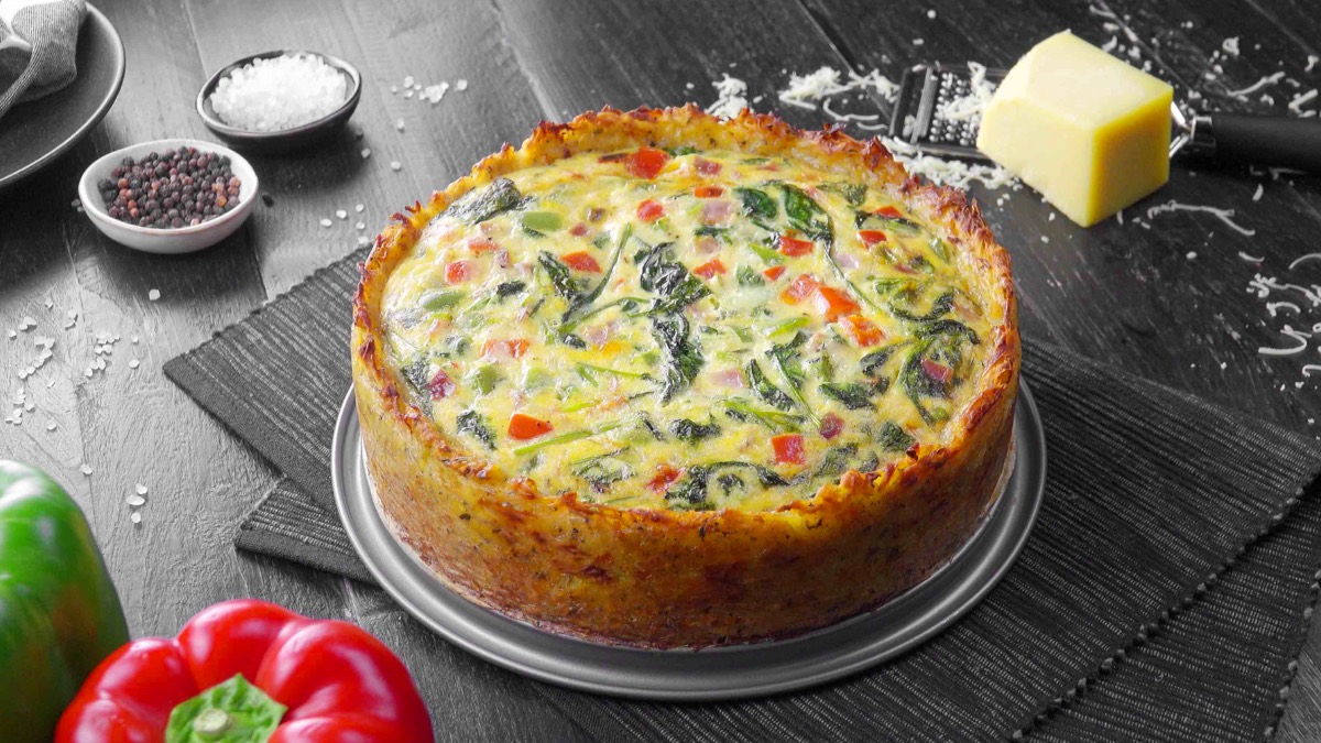 Kartoffel-Quiche auf einem Teller, darunter liegen zwei Tischsets. Drum herum liegen Paprika, Butter, Salz und Pfeffer als Dekoration.