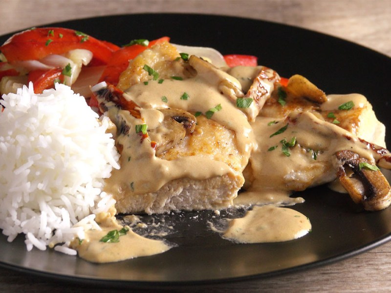 Eine Portion Parmesanhühnchen mit cremiger Champignon-Tomatensoße und Reis auf einem schwarzen Teller.