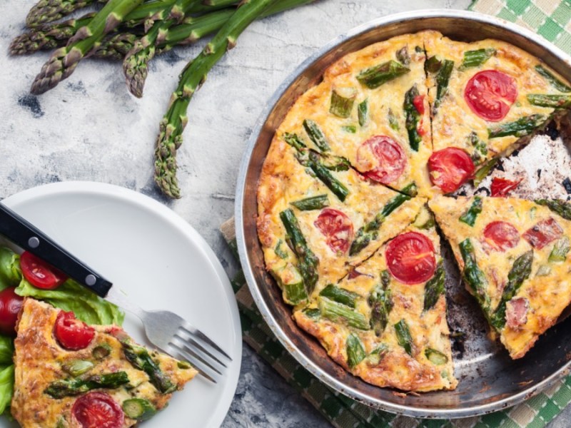 Eine spanische Tortilla mit grünem Spargel und Tomaten in der Draufsicht. Daneben steht ein Teller mit einem Stück Tortilla.