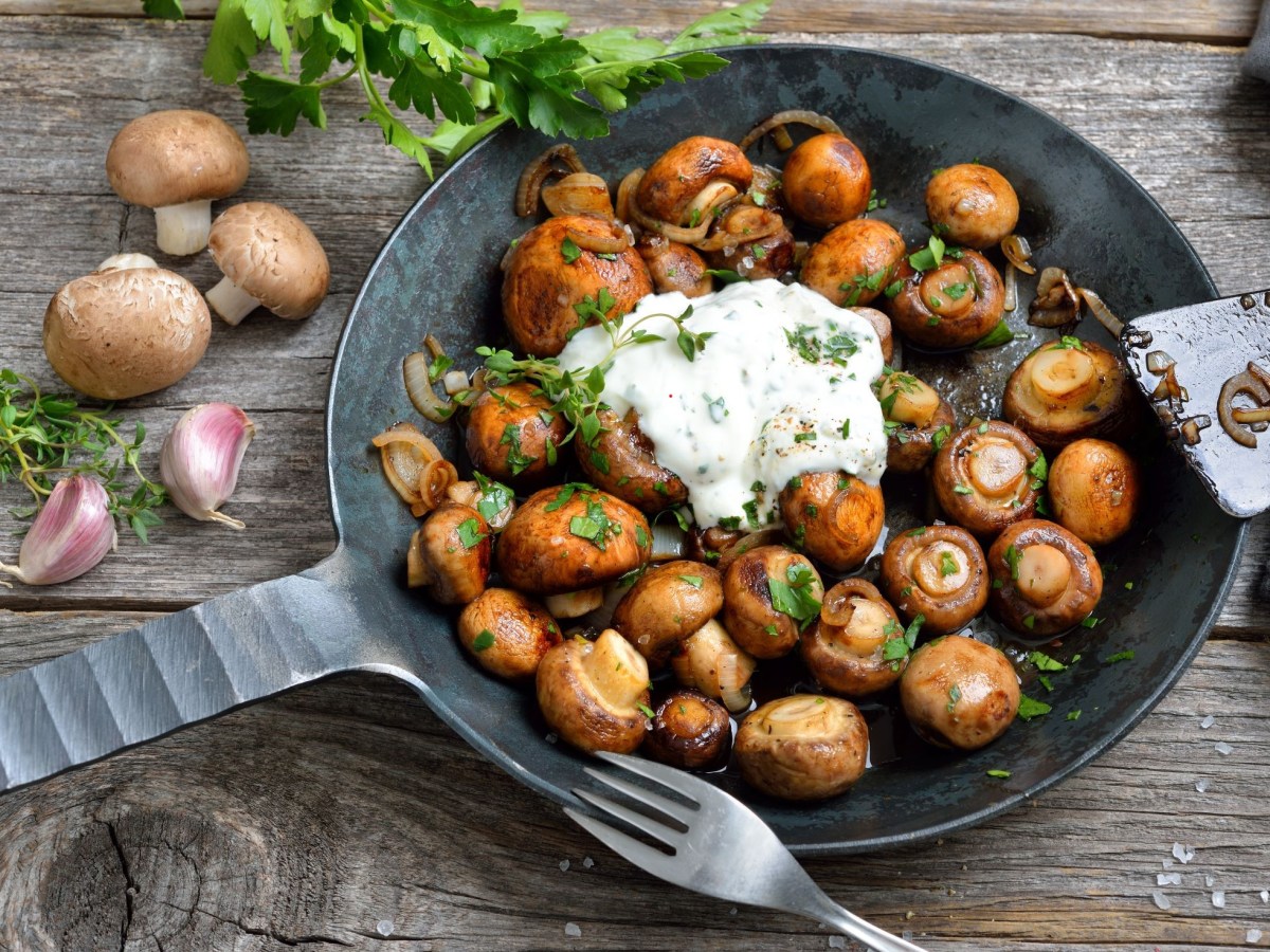 Gusseiserne Pfanne gefüllt mit Champignons wie vom Weihnachtsmarkt. Ein Klecks Knoblauchsoße in der Mitte. Pilze und Kräuter als Deko im Hintergrund.