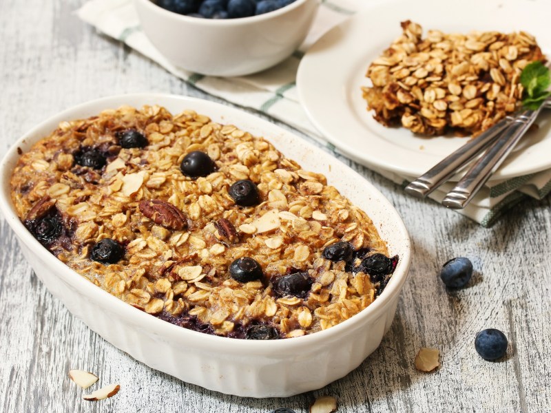 Eine Auflaufform mit gebackenen Blaubeer-Bananen-Haferflocken und Nüssen, daneben ein Teller mit einer Portion.