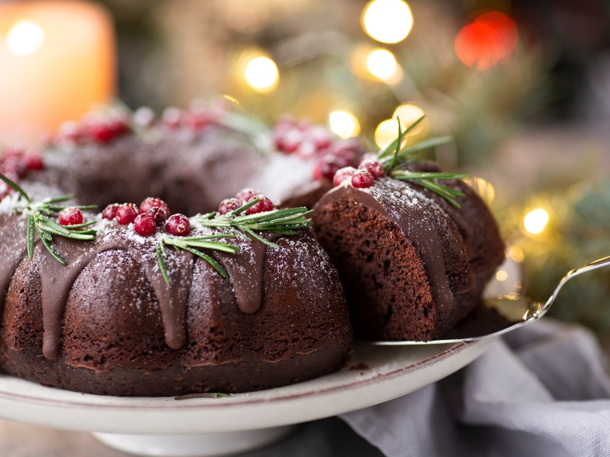 Ein angeschnittener Glühwein-Gugelhupf, von dem mit einem Tortenheber ein Stück herausgenommen wird, im Hintergrund unscharf Lichterketten.