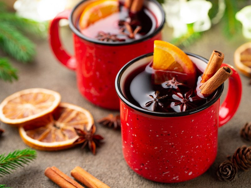 2 rote Tasse mit Glühwein, der mit Glühweingewürzen wie Zimt und Anis sowie Orangenscheiben garniert ist.