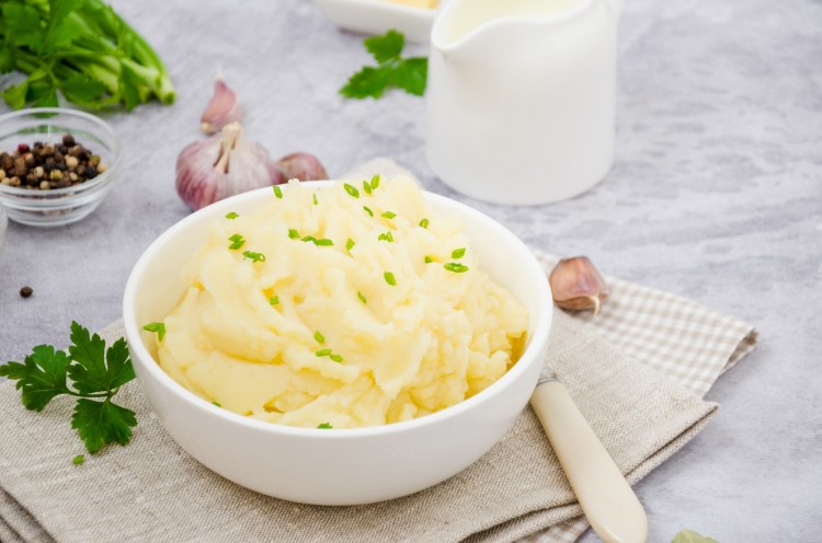 Eine weiße Schüssel Knoblauch-Kartoffelpüree, daneben ein Löffel, Knoblauchzehen und Petersilie sowie ein Milchkännchen.