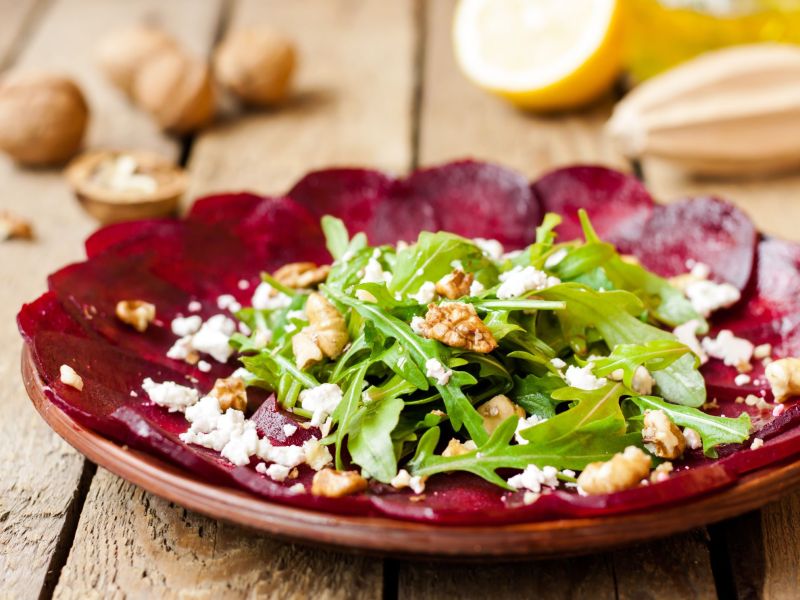 Rote-Bete-Carpaccio mit Schafskäse, Rucola und Walnüssen, serviert auf einem rot-braunen Teller. Im Hintergrund sind Walnüsse, Zitronen und eine Zitronenpresse zu sehen.