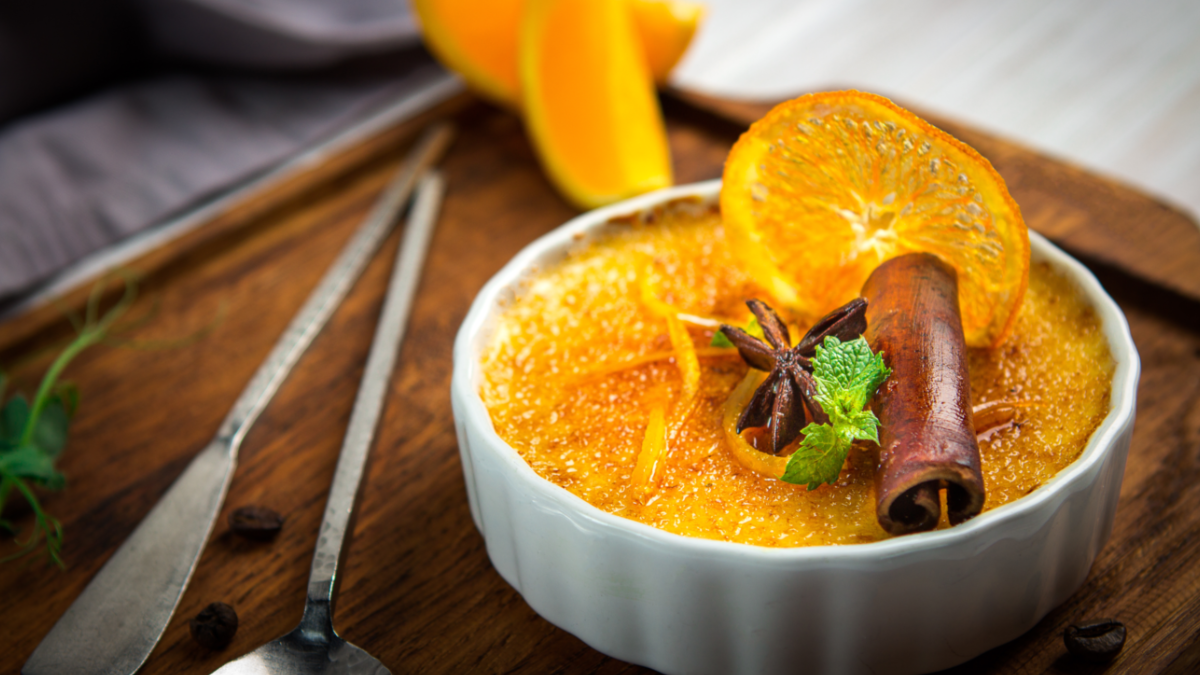 Ein überbackener Orangenpudding mit Minze, Zimtstange und Orangenscheibe in Schälchen auf Holzbrett, im Hintergrund eine angeschnittene Orange.