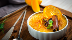 Ein überbackener Orangenpudding mit Minze, Zimtstange und Orangenscheibe in Schälchen auf Holzbrett, im Hintergrund eine angeschnittene Orange.