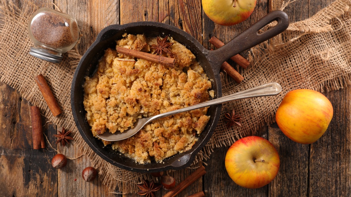 Eine Pfanne mit Apfel-Stollen-Crumble in der Draufsicht. Daneben liegen Äpfel und Zimtstange. Im Crumble liegt ein Servierlöffel.