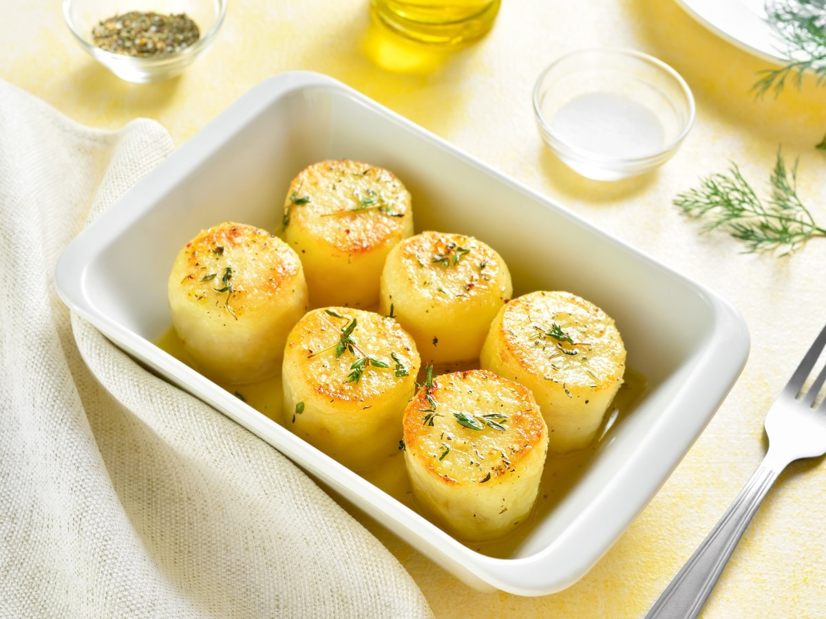 Eine weiße rechteckige Auflaufform mit Fondant-Kartoffeln, bestreut mit Rosmarin, daneben eine Gabel und ein paar Zutaten wie Brühe und flüssige Butter.