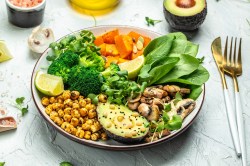 Eine herzhafte Frühstücksbowl mit Spinat, Avocado, Pilzen, Kichererbsen, Brokkoli und Süßkartoffel, daneben Gabel und Messer, außerdem Zutaten.