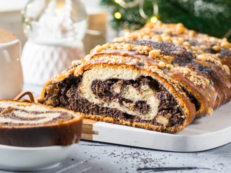 Ein aufgeschnittener Mohnstriezel mit Walnüssen und Streuseln, der auf einer länglichen Servierplatte liegt. Links davor liegt ein Stück auf einem kleinen Teller. Im Hintergrund sind ein Milchkännchen, eine Schneekugel und Tannengrün mit einer Lichterkette zu sehen.