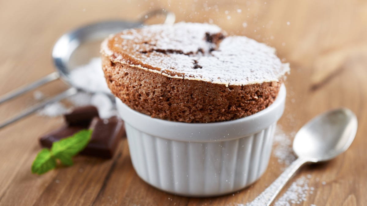 Ein schokoladensoufflé in einer kleinen Form, das mit Puderzucker bestreut wurde. Daneben ein Löffel, einige Stücke Schokolade, ein Sieb und frische Minze.