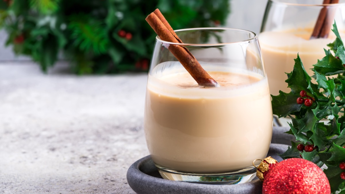 Ein Glas mit Spekulatius-Likör, in dem eine Zimtstange steckt. Im Hintergrund steht ein weiteres Glas, einige Zweige Stechpalme und eine Christbaumkugel.