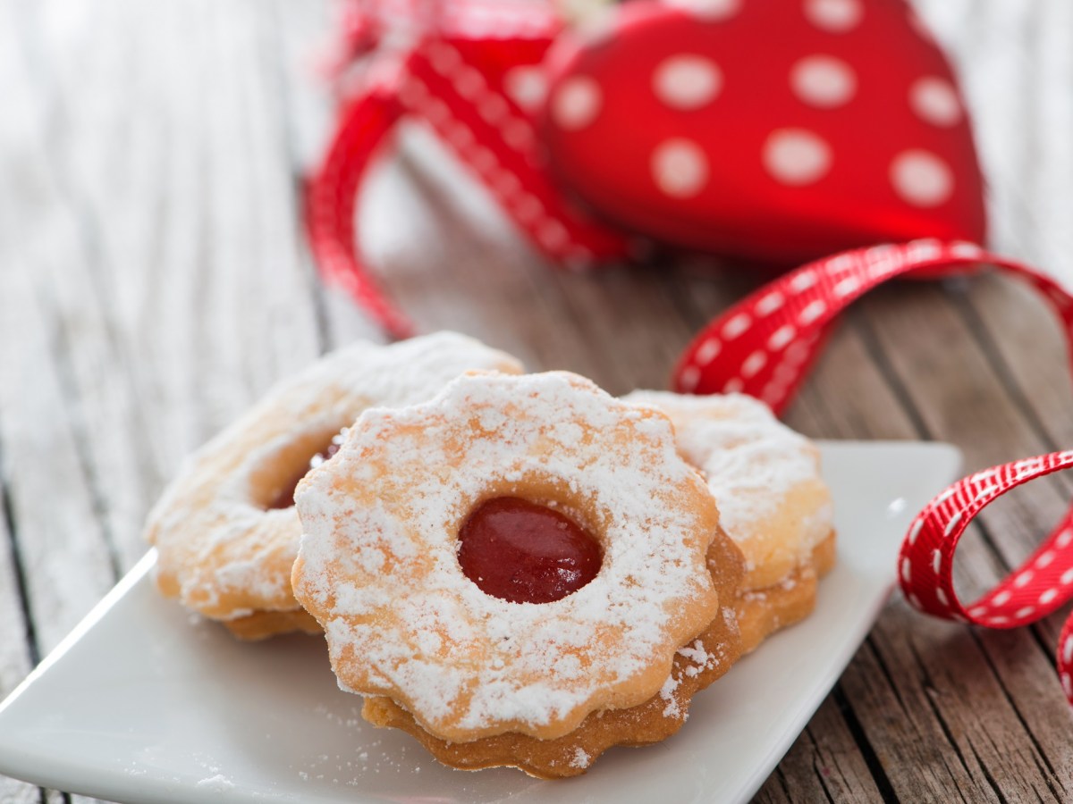 3 Spitzbuben-Plätzchen auf einem Teller, daneben weihnachtliches rotes Geschenkband.