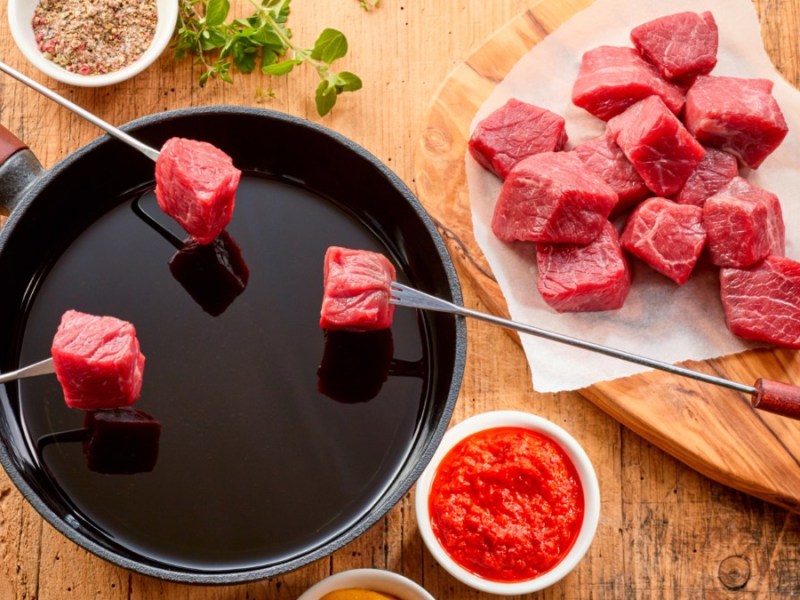 Ein Fondue, übder das 3 Fonduegabeln mit Fleisch gehalten werden, in der Draufsicht. Daneben Schälchen mit Soßen und ein Teller voller Fleisch.