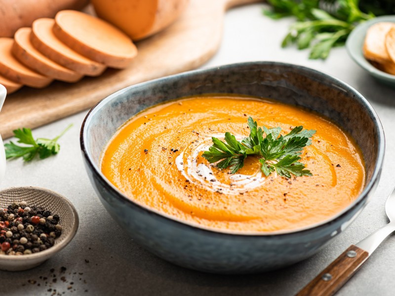 Eine Schüssel Winterliche Süßkartoffelsuppe mit Linsen und Zimt, garniert mit Petersilie und einem Klecks Schmand, daneben Süßkartoffeln geschnitten in Scheiben.