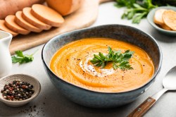 Eine Schüssel Winterliche Süßkartoffelsuppe mit Linsen und Zimt, garniert mit Petersilie und einem Klecks Schmand, daneben Süßkartoffeln geschnitten in Scheiben.