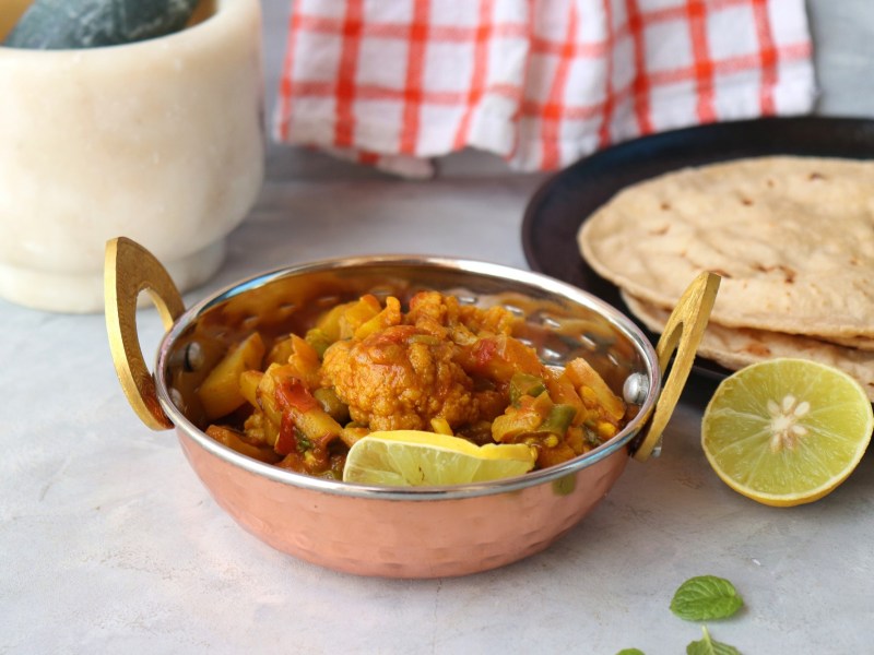 Eine Schüssel Aloo Gobi mit Blumenkohl und Kartoffeln, garniert mit einer Zitronenscheibe, daneben ein Teller mit Fladenbrot.