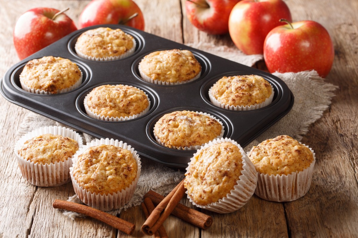 Ein Muffinblech mit Apfelmus-Muffins, drumherum weitere Muffins und frische Äpfel sowie Zimtstangen.