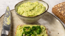 Ein großer Teller mit einer Schale Avocado-Cashew-Aufstrich, einer Scheibe Brot mit Aufstrich und einem Messer. Im Hintergrund liegt ein Vollkornbrötchen.