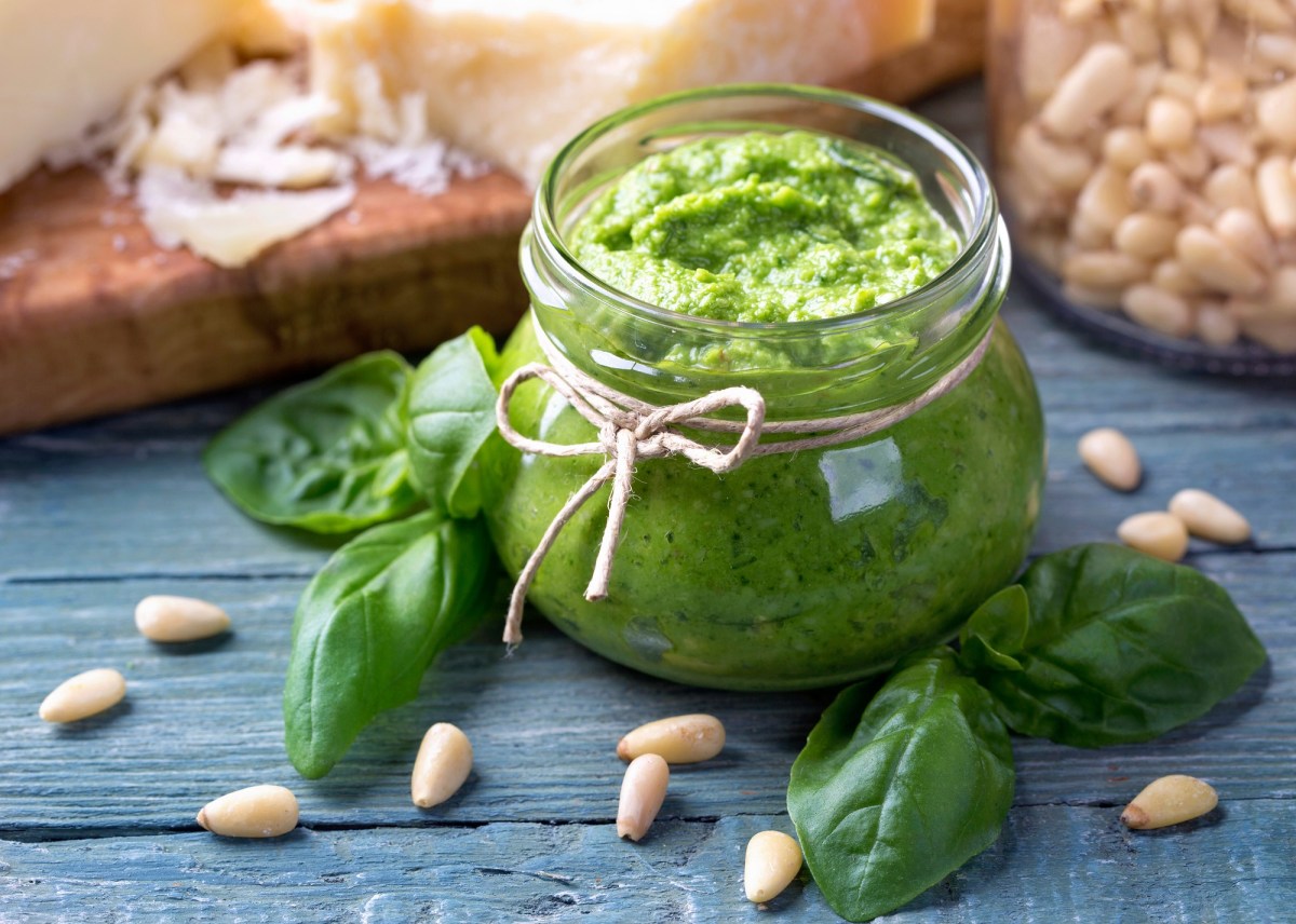 Ein Glas Basilikumpesto verziert mit einer Schleife, darum herum Basilikumblätter und Pinienkerne.