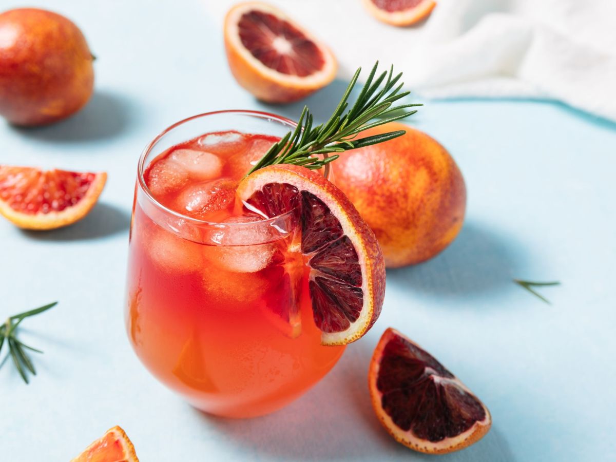 Blutorangen Rosé Spritz, serviert mit einer Scheibe Blutorange und einem Rosmarinzweig. Einige Scheiben Blutorange liegen als Deko im Hintergrund.