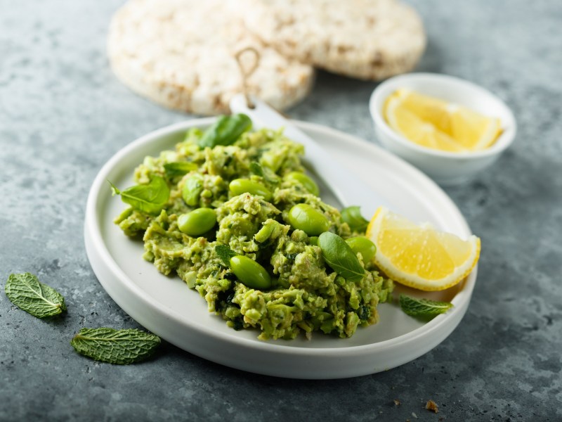 Edamame-Guacamole, mit etwas Minze garniert, auf einem weißen Teller. Serviert mit Pita-Brot.