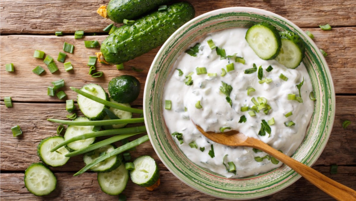 Eine Schüssel mit Gurken-Raita, in dem ein Holzlöffel liegt. Daneben liegen Gurken und frische Zutaten.