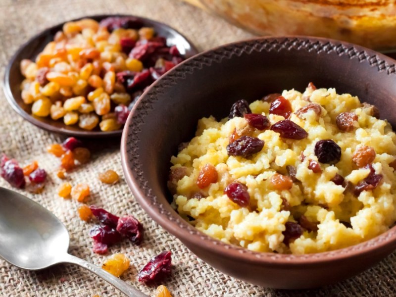 Eine Schüssel mit süßen Hirsebrei mit Rosinen und Cranberrys. Daneben liegt ein Löffel und eine kleine Schale mit Rosinen.