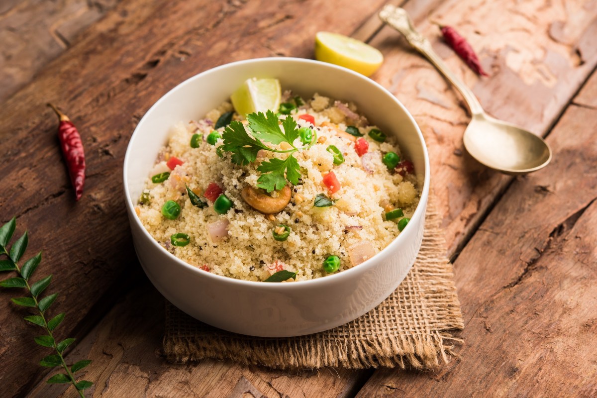 Eine Schale indisches Upma, Hartweizengrieß, mit etwas Gemüse und Kräutern, daneben ein Löffel.