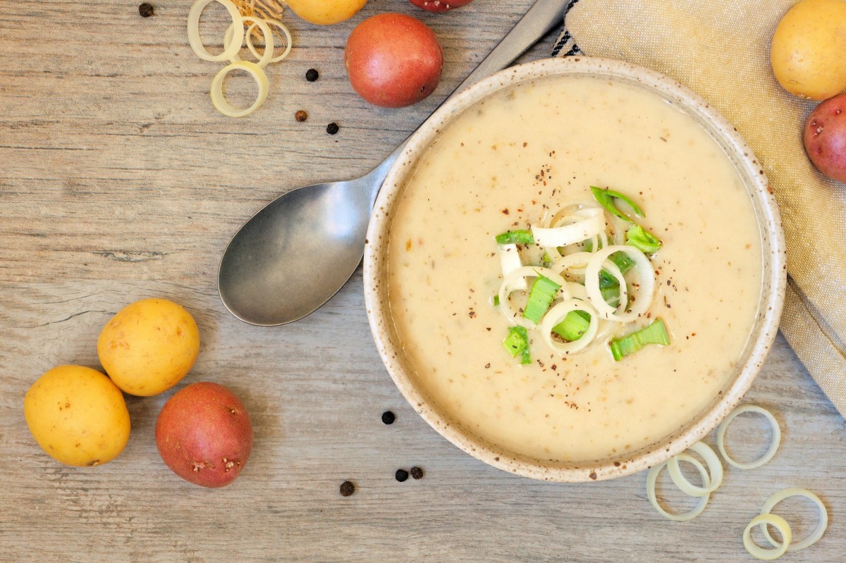 Eine Schüssel Kartoffel-Lauch-Suppe mit Lauch und Pfeffer garniert, daneben ein Löffel und Kartoffeln, Draufsicht.