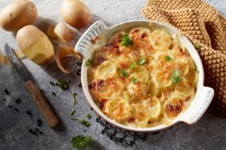 Eine runde Auflaufform mit Kartoffelgratin mit Pilzen, daneben teilweise geschälte Kartoffeln und ein Messer sowie Kräuter.