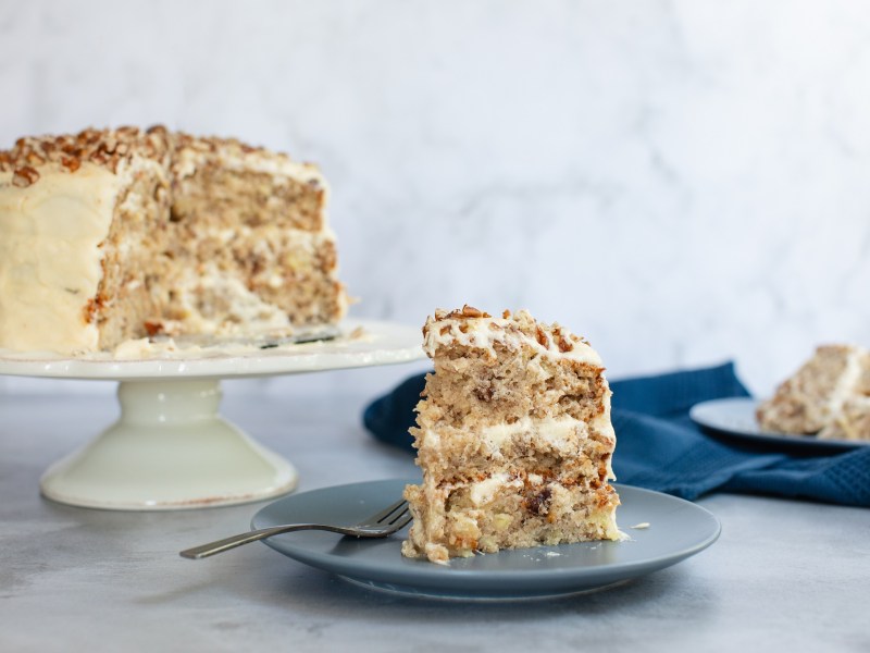 Ein Stück Kolibri-Kuchen auf einem Teller, im Hintergrund der angeschnittene Kuchen mit weißer Glasur verziert.