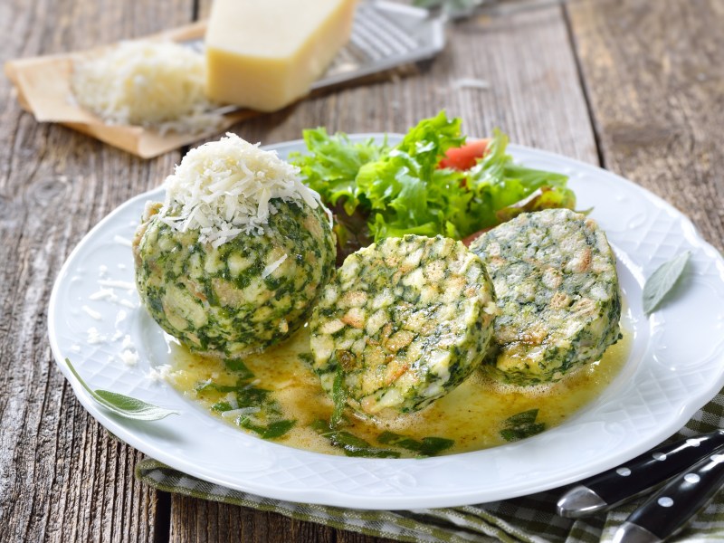 2 Spinatknödel, einer halbiert, auf einem Teller mit Salat, im Hintergrund ein Stück Parmesan, neben dem Teller Besteck.