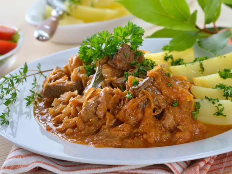 Eine Portion Szegediner Gulasch mit Kartoffeln auf einem weißen Teller. Im Hintergrund stehen eine Schüssel mit Kartoffeln und eine mit Salat.