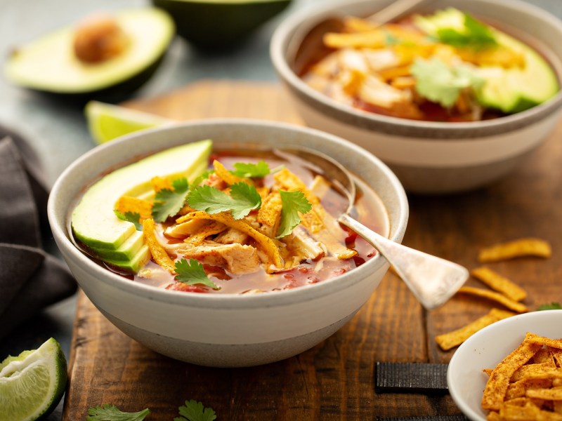 Zwei Schüsseln Tortilla-Suppe mit Avocado und Tortilla-Streifen, drumherum verschiedene Zutaten wie eine halbierte Avocado.