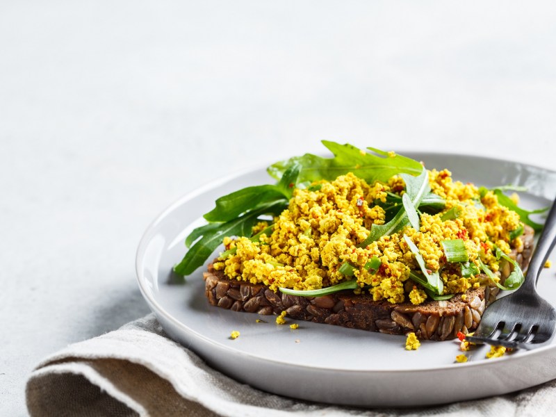 Ein Brot belegt mit veganem Rührei und Rucola auf einem Teller, Nahaufnahme.