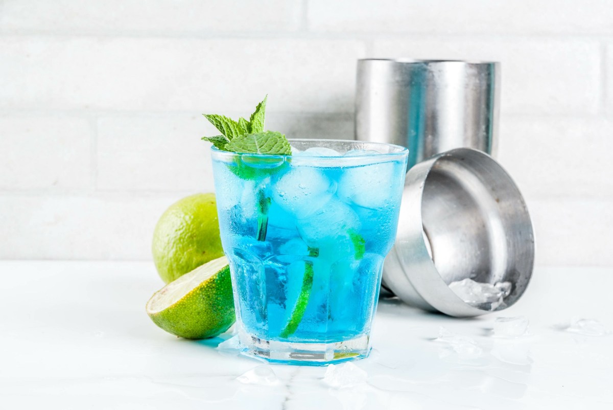 Ein Glas Blue Lagoon mit Eiswürfeln, Limettenscheiben und Minze garniert, dahinter ein Cocktailshaker und halbierte Limetten.