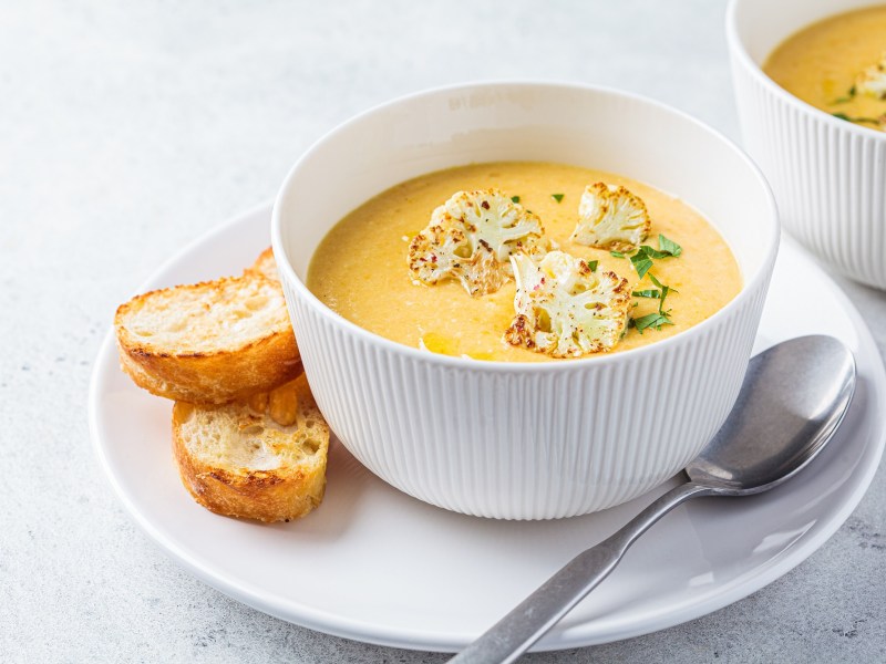 Eine Schüssel Blumenkohl-Kartoffel-Suppe mit geröstetem Blumenkohl garniert auf einem Teller, auf dem auch ein Löffel und zwei Scheiben getoastetes Weißbrot liegen.