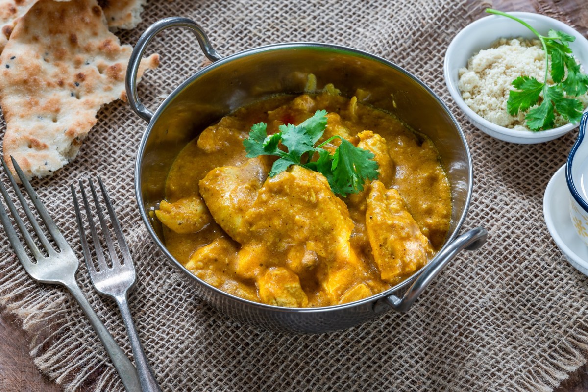 Chicken Korma in einer Metallschale mit Koriander garniert. Daneben liegen Naan-Brot, zwei Gabeln und eine Schale gemahlene Mandeln.